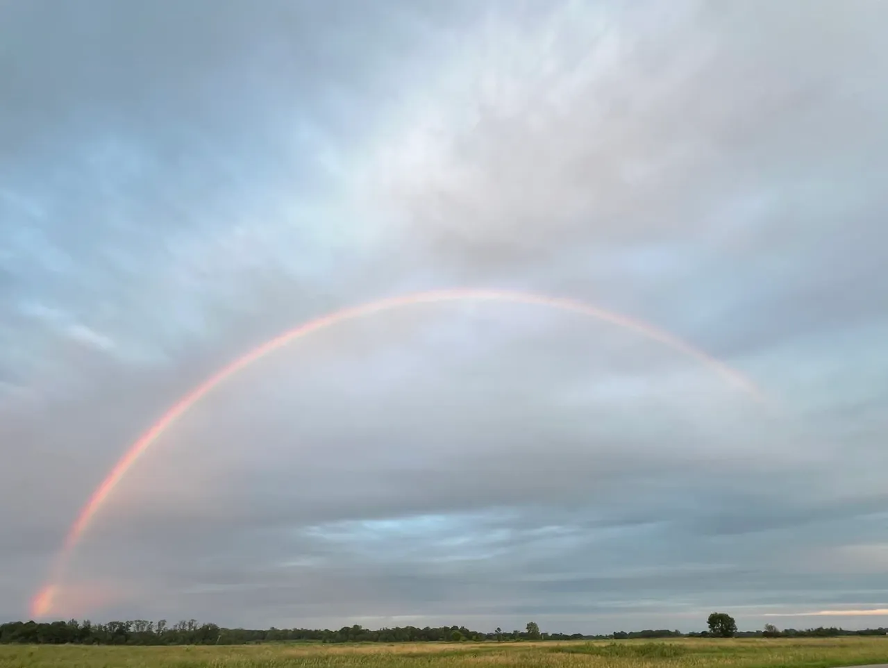 A faint rainbow arches over a cloudy sky above a green field.